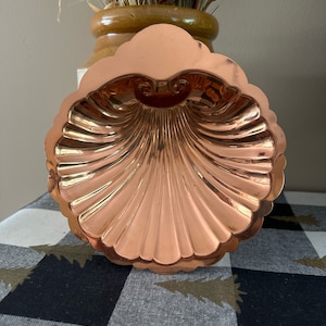 May include: A copper shell-shaped dish with a fluted design. The dish is shiny and has a warm, metallic color.
