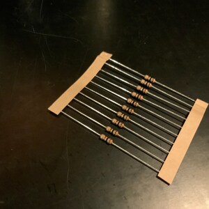 May include: A close-up of a strip of brown paper with ten silver resistors attached to it. The resistors are arranged in a row, with the brown paper strip running along the top and bottom of the resistors.