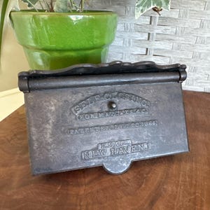 May include: A vintage, rectangular, cast iron match safe with the text "SELF CLOSING FOR MATCHES & C." and "PATENTED. DEC. 20. 1864." The safe is dark gray and sits on a wooden surface. The text "D.M.& Co. NEW HAVEN" is also visible.