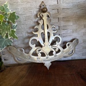 May include: A brass inclinometer featuring an ornate, scrollwork frame with a central vertical pointer. The curved base displays a graduated scale marked with numbers ranging from 0 to 30 on both sides, designed for measuring angles or tilt.