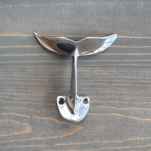 May include: A silver whale tail shaped hook. The hook is mounted on a wooden surface.