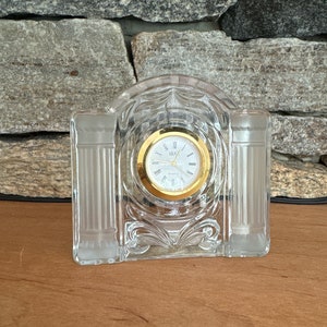 May include: A clear crystal clock with a gold face and Roman numeral markings. The clock is shaped like a small archway with pillars on either side.