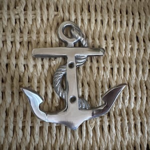 May include: A silver-colored anchor with rope detailing. The anchor is set against a woven, beige background. The anchor's design includes a ring, a crossbar, and two flukes. The rope is wrapped around the anchor's shank.