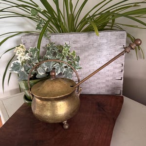 May include: A brass pot with a rounded body, three small feet, and a matching lid. It features a curved metal handle attached to the sides and a long, slender metal spoon resting inside the pot through a small notch in the lid's edge.