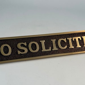 May include: A rectangular bronze-colored sign with the words "NO SOLICITING" in raised gold lettering. The sign has a textured background and decorative edges, with a hole on each end for mounting.