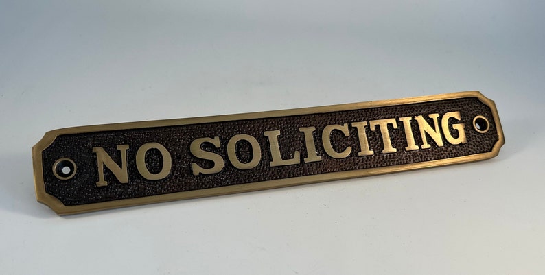 May include: A rectangular bronze-colored sign with the words "NO SOLICITING" in raised gold lettering. The sign has a textured background and decorative edges, with a hole on each end for mounting.