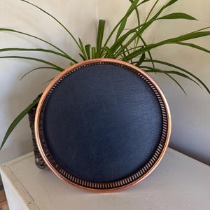 May include: A round tray with a copper rim and a black leather bottom. The tray is sitting on a white surface.