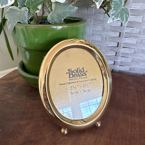 May include: An oval-shaped solid brass photo frame with a polished finish and two small ball feet. The glass insert features text reading Solid Brass Photo Frame Hand Polished & Lacquer Coated 2 1/2 inches by 3 1/2 inches, 6 cm by 9 cm.