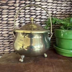 May include: A small, antique brass pot with a lid and handle. The pot has three small ball feet and a shiny, reflective surface. The handle arches over the top, and the lid has a small knob. The pot is likely used for decorative purposes.