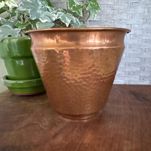 May include: A hammered copper planter with a wide rim and a textured surface. The planter is empty and sits on a wooden surface. A green ceramic pot with a plant is visible in the background.