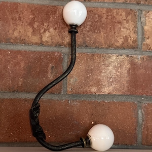 May include: A black metal hook with two white ceramic knobs. The hook is mounted on a brick wall.