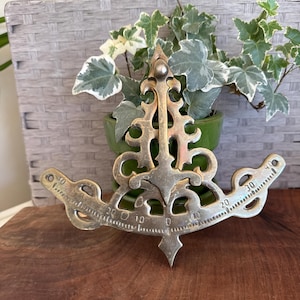 May include: An antique brass instrument with a decorative, ornate design. The instrument has a curved shape with a central vertical element and two horizontal arms, each marked with numbers from 0 to 40. The background includes a green potted plant and a wooden surface.