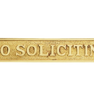 May include: A rectangular brass sign with the words "NO SOLICITING" in raised lettering. The sign has a textured background and decorative edges, with holes for mounting.