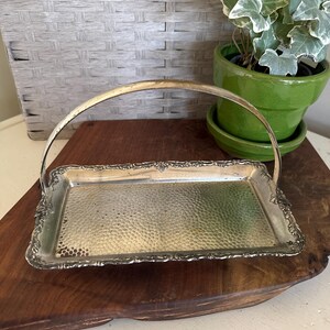 May include: A vintage silver-plated serving tray with a hammered surface and ornate edges. The tray has a curved handle and sits on a wooden surface. A green potted plant is in the background.
