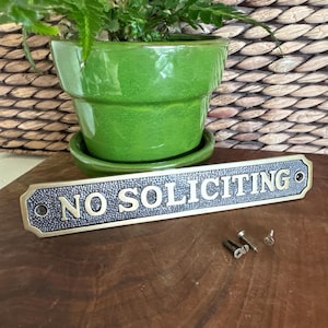 May include: A green ceramic plant pot with a matching saucer holds a leafy green plant. A rectangular brass sign with the words "NO SOLICITING" in gold lettering is in front of the pot. Small screws are next to the sign.