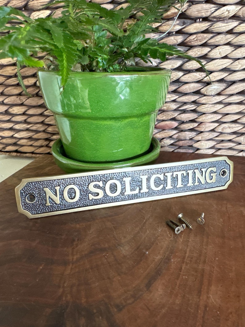 May include: A green ceramic plant pot with a matching saucer holds a leafy green plant. A rectangular brass sign with the words "NO SOLICITING" in gold lettering is in front of the pot. Small screws are next to the sign.