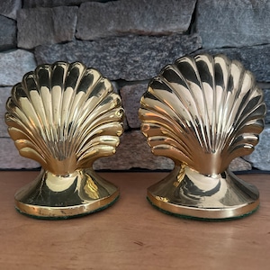 May include: A pair of gold-tone brass bookends shaped like seashells. The bookends are detailed with a raised, textured design.