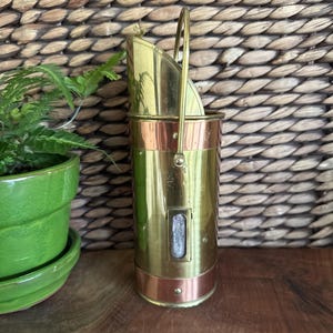 May include: A vintage brass and copper coal scuttle with a handle. The cylindrical body is brass with copper bands at the top and bottom. A small rectangular window is visible on the front. The scuttle is set against a woven background.