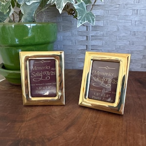 May include: Two rectangular gold-colored brass photo frames. Each frame has a polished finish and features the text "Memories in Solid Bras" and "1.5" x 2.5" Photo Frame". The frames are displayed on a wooden surface.