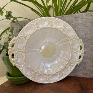 May include: A white, decorative serving plate with a seashell design around the rim. The plate has a scalloped edge and two handles. The center of the plate has a textured, radial pattern.