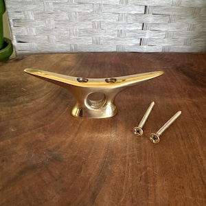 May include: A polished brass boat cleat with a smooth, curved design and two pre-drilled mounting holes. The cleat is accompanied by two matching brass screws with Phillips heads, positioned to the right of the hardware on a wooden surface.