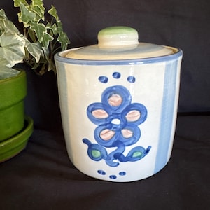 May include: A cylindrical ceramic canister with a lid, featuring a hand-painted blue floral motif on a cream-colored background. The design includes light blue vertical stripes on the sides and a small green knob on the center of the lid.