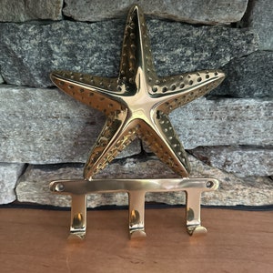 May include: A brass starfish key holder with three hooks. The starfish is detailed with small holes and is mounted above a horizontal bar with three curved hooks. The key holder is set against a stone wall.