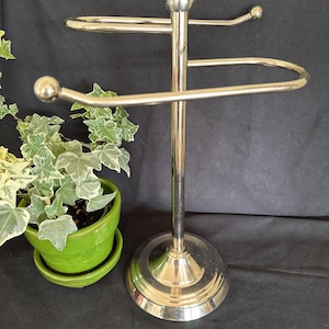 May include: A silver-toned metal towel stand featuring a central vertical pole, a circular base, and two horizontal arms with spherical finials at the ends. The stand is constructed with a polished finish and a sturdy, weighted base design.