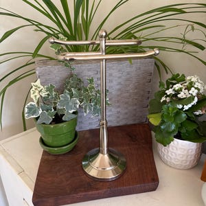 May include: A polished silver-tone metal countertop towel stand featuring a central vertical post with a circular base and two horizontal arms extending in opposite directions, each finished with a small spherical cap at the end.