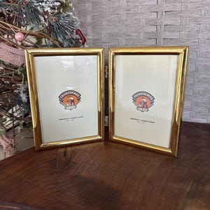 May include: Two gold-colored picture frames with a rectangular shape. Each frame has a cream-colored interior with the "Frameworks USA" logo. The frames are made of solid brass with a lacquer coating. The frames are 4 x 6 inches.