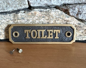 May include: A brass sign with the word "TOILET" embossed in black lettering. The sign has two small holes on either side for mounting. Two screws are included.