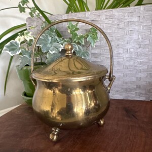 May include: A vintage brass pot with a lid and handle. The pot has a rounded body, three small feet, and a curved handle. The brass has a warm, golden color and reflects light. The pot is likely used for decorative purposes.