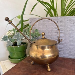 May include: A vintage brass sugar bowl with a lid and a handle. The bowl has three small feet and a matching spoon. The brass has a warm, aged patina. The bowl is sitting on a wooden surface.