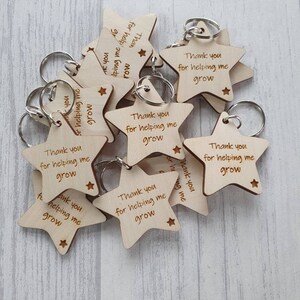 Peut inclure: Un ensemble de porte-cl&eacute;s en bois en forme d'&eacute;toile avec le texte "Merci de m'aider &agrave; grandir" grav&eacute; sur chacun. Les porte-cl&eacute;s sont en bois brun clair et ont des anneaux de cl&eacute;s argent&eacute;s.