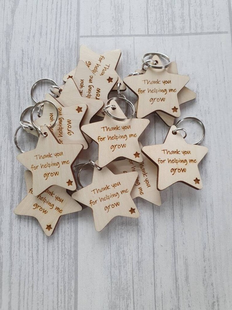 Peut inclure: Un ensemble de porte-cl&eacute;s en bois en forme d'&eacute;toile avec le texte "Merci de m'aider &agrave; grandir" grav&eacute; sur chacun. Les porte-cl&eacute;s sont en bois brun clair et ont des anneaux de cl&eacute;s argent&eacute;s.