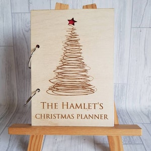 May include: A wooden Christmas planner with a laser-cut design of a Christmas tree with a red star on top. The planner is titled "The Hamlet's Christmas Planner".