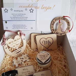 May include: A white gift box filled with wedding-themed decorations, including a wooden horseshoe, a wooden heart ornament, a wooden puzzle piece, a wooden sign with the text "Mr & Mrs Green Est. 09.08.19", a candle with the text "To the beautiful Bride on her wedding day Mrs Green 09 August 2019", and a red and white wooden ornament with the text "Our First Christmas as Mr & Mrs Green".