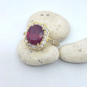Pode incluir: Um anel de ouro com uma grande gema vermelha de corte almofada, cercada por um halo de gemas brancas menores.