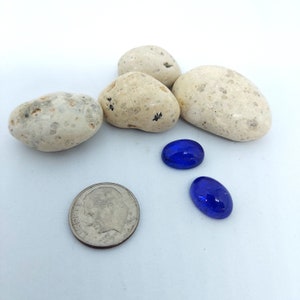 Puede incluir: Cuatro piedras blancas moteadas y dos piedras de vidrio ovaladas azules sobre una superficie blanca.
