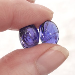 Puede incluir: Dos piedras preciosas ovaladas, transl&uacute;cidas, de color p&uacute;rpura con un acabado brillante e iridiscente.