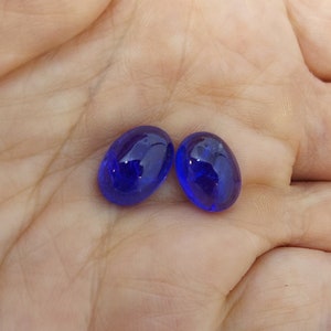 Puede incluir: Dos piedras preciosas azules de forma ovalada, probablemente zafiros, se muestran en una mano. Las piedras preciosas son lisas y tienen un acabado brillante.