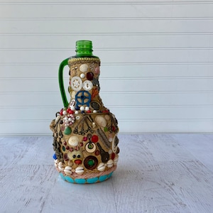 May include: A decorative bottle with a green handle and neck, adorned with various embellishments. The bottle is covered in seashells, buttons, beads, and other small decorative items. The base is a light blue color, and the bottle is set against a white background.