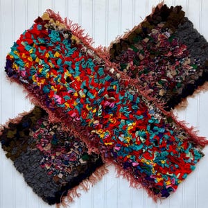 May include: Two handmade rag rugs in a cross shape. The rugs feature a patchwork design with vibrant colors including red, turquoise, and pink. The edges are finished with a fringed trim. The rugs are placed on a white wooden surface.
