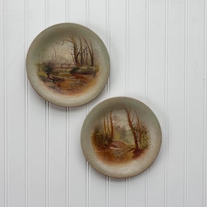 May include: Two round decorative plates with landscape paintings. Each plate features a serene scene of a river, trees, and a bridge, painted in muted tones of brown, green, and beige. The plates are displayed against a white background.