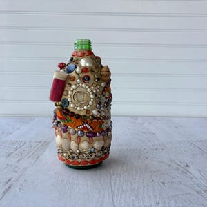 Peut inclure: Une bouteille décorative avec un bouchon vert, ornée de divers ornements. La bouteille est recouverte de coquillages, de perles et d'autres éléments décoratifs. Un petit objet rouge est attaché sur le côté. La bouteille est une œuvre d'art unique.