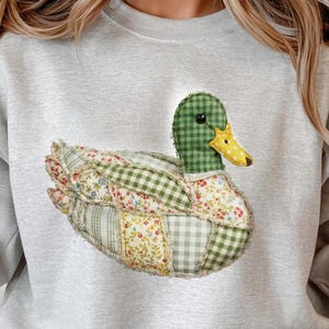 May include: A light gray sweatshirt featuring a patchwork duck design. The duck has a green checkered head, a yellow and white polka-dotted beak, and a body made of floral and checkered fabric patches. The sweatshirt has long sleeves.