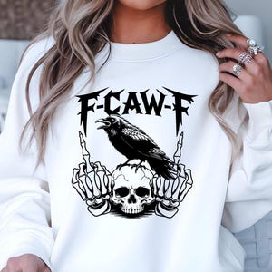 May include: White sweatshirt with a black graphic featuring a raven perched on a skull, with skeleton hands giving the middle finger. The text "F-CAW-F" is above the raven. A casual, edgy design.