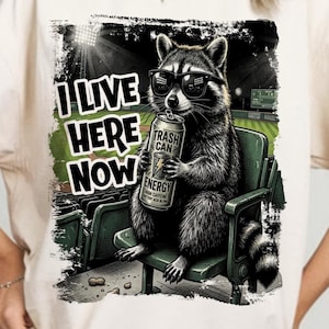 Può includere: T-shirt color crema con un procione con occhiali da sole, che tiene in mano una lattina di "Trash Can Energy" in uno stadio. Il testo sulla maglietta recita "I Live Here Now."