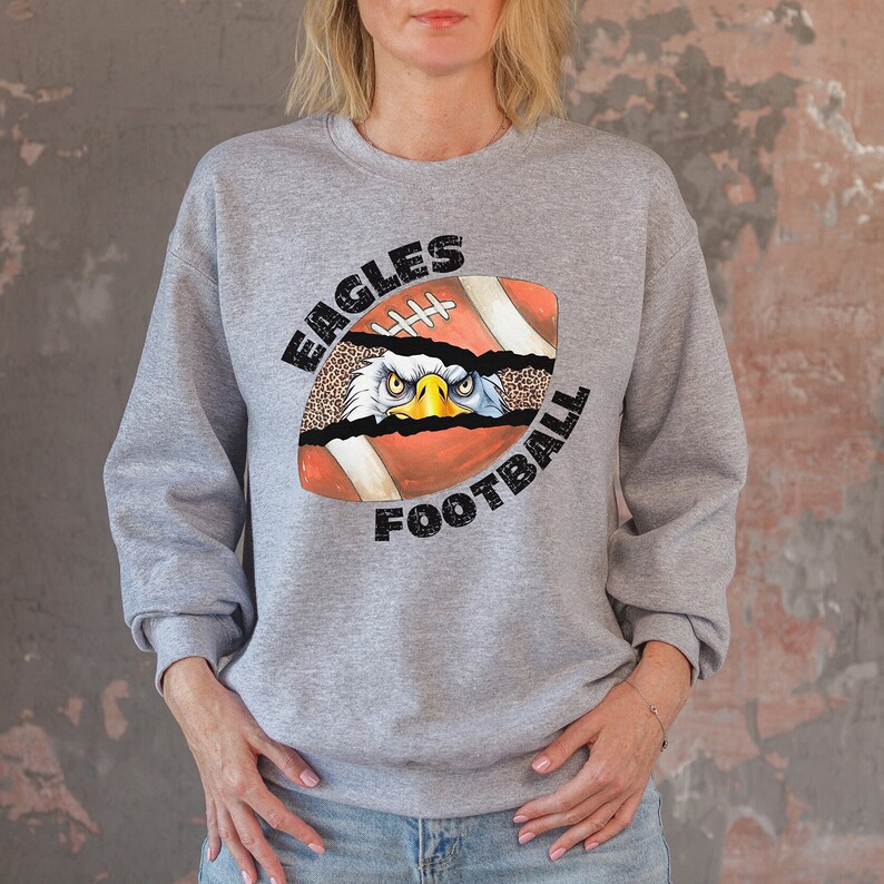 Pode incluir: Camisola cinzenta com um gr&aacute;fico de uma bola de futebol com a cabe&ccedil;a de uma &aacute;guia a espreitar do meio. O texto "EAGLES FOOTBALL" est&aacute; impresso em torno da bola de futebol.