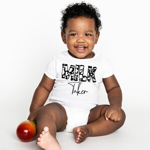 May include: A white baby onesie with a black and white graphic of the word "MLK" and the text "Taken" underneath. A baby is wearing the onesie and sitting on a white surface holding a green and orange toy.